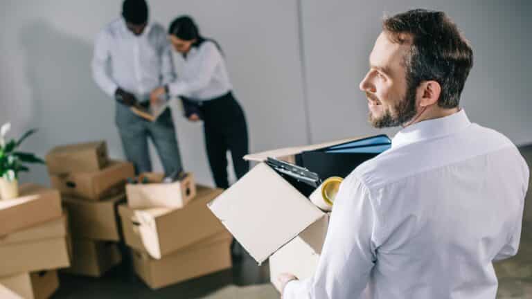 back view of businessman holding cardboard box with office supplies while colleagues unpacking boxes in new office